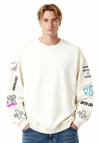 Krémová mikina s dlhými rukávmi s grafickými potlačami v ružovej, čiernej a modrej farbe. Hladký golier a manžety; voľný strih.