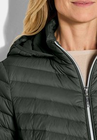 Donkergroene pufferjack met horizontale stitching, capuchon en een zilveren rits aan de voorkant. De stof lijkt glad en lichtgewicht te zijn.
