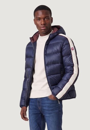 Mand iført en blå dunjakke med hvid og rød stribe på ærmet, hvid sweater og blå jeans, står med den ene hånd i lommen.
