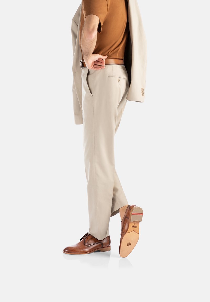 Chaussures en cuir marron à lacets, pantalon beige et haut marron. Stylisé avec un blazer de couleur claire posé sur une épaule.