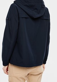 Chaqueta ligera azul marino con capucha, puños elásticos y dobladillo con cordón ajustable. Presenta una textura sutil y un pequeño logotipo en la manga.