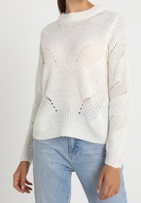 Maglione bianco lavorato a maglia con taglio morbido, caratterizzato da un collo a costine e un motivo traforato, abbinato a jeans in denim blu chiaro.