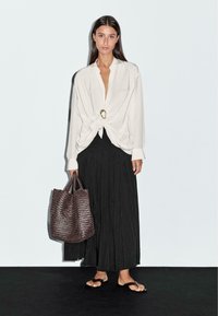 Donna che indossa una blusa drappeggiata bianca con un dettaglio ad anello dorato, una gonna plissettata nera, sandali neri e che porta una grande borsa a tracolla intrecciata marrone.