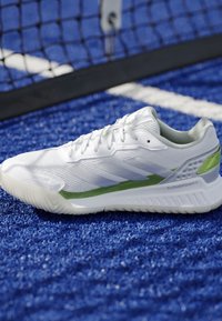 Scarpa da atletica bianca con accenti verdi su un campo sportivo blu testurizzato vicino a una rete nera.