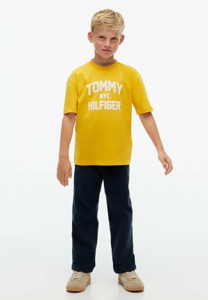 Lyshåret dreng står med arme afslappet, iført en gul Tommy Hilfiger t-shirt, mørke bukser og lyse beige sneakers på en hvid baggrund.