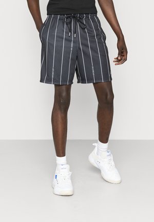 Mann trägt schwarz-weiß gestreifte Air Jordan Shorts, weiße Socken und weiße Sportschuhe, steht mit entspannten Händen.