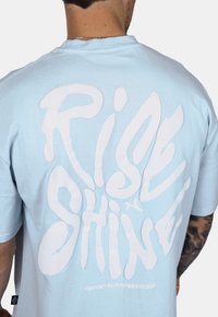 Světle modré bavlněné tričko s velkým bílým grafickým potiskem, který na zádech nese nápis "Rise & Shine", s volným střihem a krátkými rukávy.