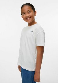 Camiseta de algodón blanca con mangas cortas, cuello redondo y una pequeña impresión negra de "DESAYUNO" en el lado izquierdo. Combinada con jeans azules.