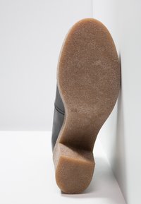 Semelle de chaussure en caoutchouc crêpe marron clair avec une texture et un talon bloc, sur un fond blanc et un mur.