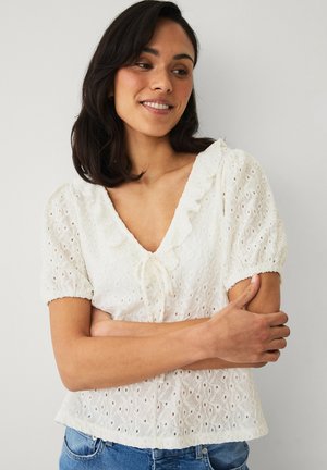 Frau mit dunklem Haar lächelt, trägt eine weiße Bluse aus Lochspitze mit Puffärmeln und gerafftem Ausschnitt, steht vor einem schlichten Hintergrund.