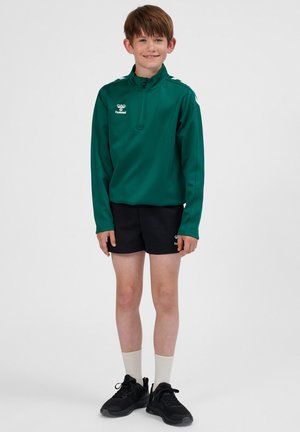 Groen jack met rits en lange mouwen, gecombineerd met zwarte shorts. Het jack heeft een logo en witte accenten op de schouders. Zwarte sportschoenen met witte sokken.
