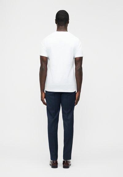 Άνδρας που στέκεται γυρισμένος προς τα πίσω, φορώντας ένα απλό λευκό κοντομάνικο μπλουζάκι, σκούρο μπλε παντελόνι και καφέ επίσημα παπούτσια, σε ουδέτερο φόντο.