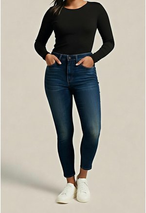 Femme portant un jean skinny bleu foncé, un haut noir à manches longues et des baskets blanches, debout les mains dans les poches.