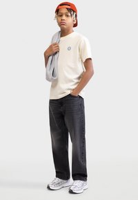 Κρεμ T-shirt με μπλε μπαλώμα με χαμογελαστό πρόσωπο, γκρι πετσέτα ιδρώτα περασμένη στον ώμο, μαύρο τζιν και ασημένια αθλητικά παπούτσια. Φορά κόκκινο καπέλο μπέιζμπολ.