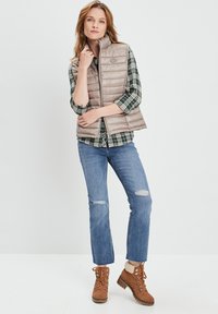 Hellbeige Steppweste mit hohem Kragen, getragen über einem grün karierter Hemd. Kombiniert mit ausgefransten blauen Jeans und braunen Schnürstiefeln.