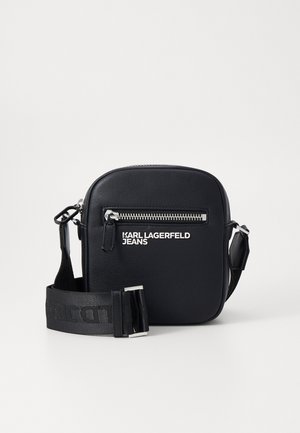 Zwarte leren schoudertas met een met rits sluitende voorvak, zilveren hardware en een verstelbare schouderband met de merknaam in vette letters.