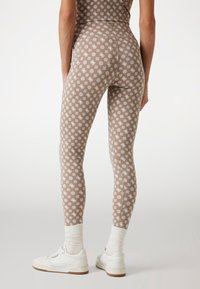 Barna leggings fehér, ismétlődő virágmintával, puha anyagból készült. Fehér sportcipővel és krémszínű zoknival párosítva.