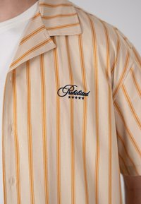 Short-sleeve shirt in beige with vertical orange stripes. Features a large navy embroidered brand name and a pointed collar.