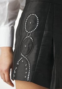 Jupe noire avec découpes circulaires décoratives et clous argentés près de l'ourlet, portée avec une chemise blanche à manches longues visible au poignet.