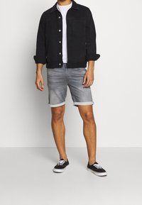 Giacca di denim nero con chiusura a bottoni, abbinata a pantaloni corti di denim grigio chiaro con risvolto e sneakers in tela nere con lacci bianchi.