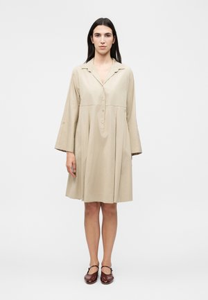 Γυναίκα που στέκεται φορώντας μια μπεζ μακριά μπλούζα, φόρεμα μήκους έως τα γόνατα με κουμπιά και καφέ παπούτσια Mary Jane μπροστά σε απλό φόντο.