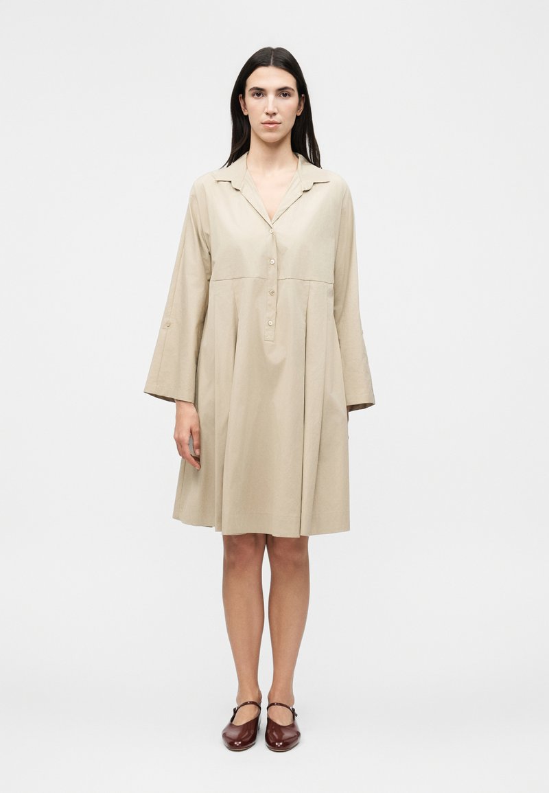 Γυναίκα που στέκεται φορώντας μια μπεζ μακριά μπλούζα, φόρεμα μήκους έως τα γόνατα με κουμπιά και καφέ παπούτσια Mary Jane μπροστά σε απλό φόντο.