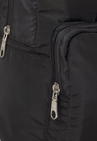 Černý polstrovaný batoh s hladkým povrchem, vybavený stříbrnými kovovými zipy a kováním. Dvojité zipové přihrádky vytvářejí funkční design.