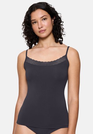 Woman with curly hair wearing a dark gray camisole with lace trim and matching underwear, standing against a plain light background.