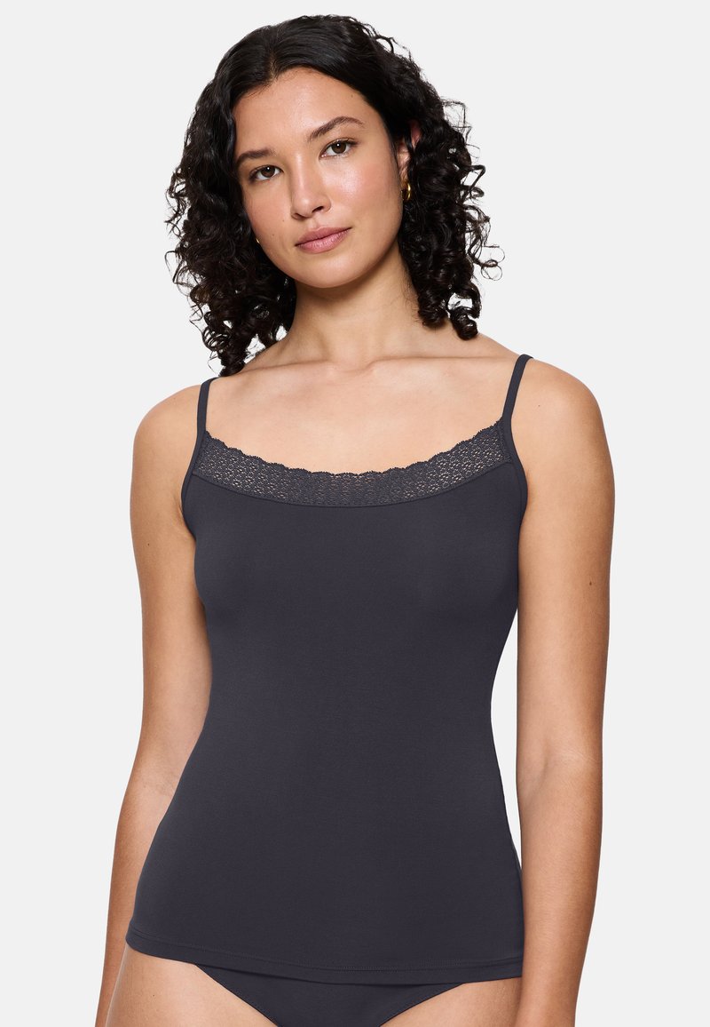 Kobieta z kręconymi włosami ubrana w ciemnoszary top na ramiączkach z koronkowym wykończeniem i dopasowane majtki, stojąca na tle gładkiego jasnego tła.