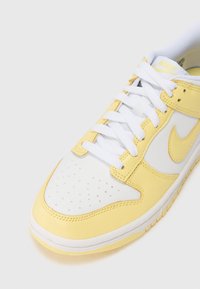 Hvite og gule sneakers med perforert tå, skinnoverdel og gummisåle. Har hvite snørebånd og Nike-logoinnslag i gult.