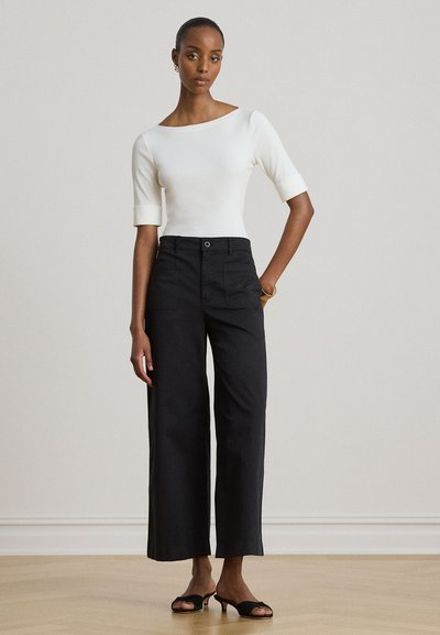 Femme portant un haut blanc ajusté avec des manches aux coudes, un pantalon noir à jambes larges et des talons ouverts noirs, debout contre un mur uni.