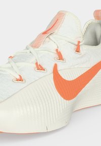 Λευκό αθλητικό παπούτσι με πορτοκαλί λογότυπο Nike και λεπτομέρειες, υφασμένο πέλμα και λευκά κορδόνια περασμένα από πορτοκαλί οφθαλμούς.