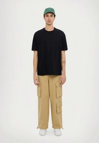 Černé tričko s krátkými rukávy a kulatým výstřihem, doplněné béžovými cargo kalhotami s několika kapsami a bílými teniskami. Na hlavě zelená kšiltovka.