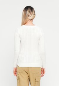 Mulher com cabelo loiro comprido vestindo um suéter branco de malha e calças cargo bege, de pé de costas contra um fundo simples.