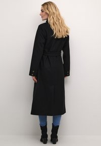 Manteau noir long avec une taille ceinturée, des poignets boutonnés et une coupe droite. Fabriqué dans un tissu texturé, avec une fente dans le dos et un design minimaliste.