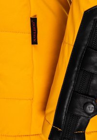Yellow quilted jacket with black leather accents. Features stitching details and a label reading "Navajo." Zipper and button hardware included.