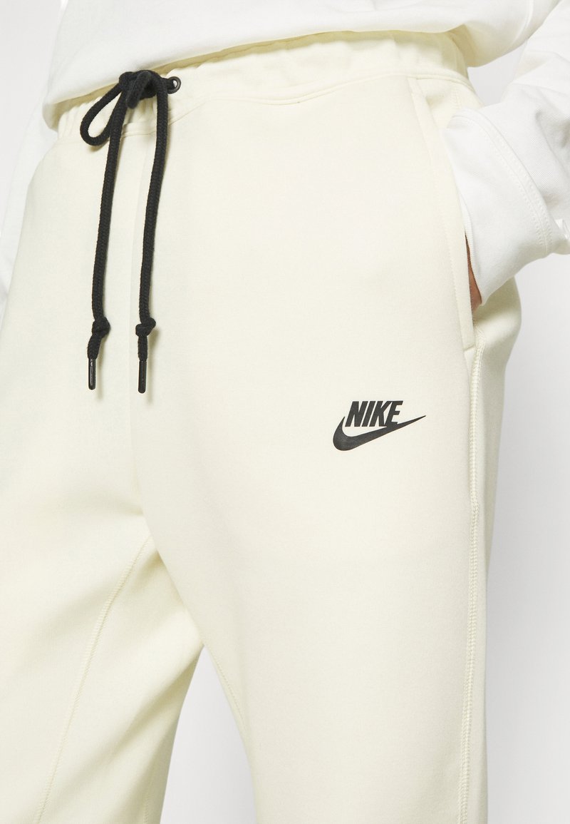 Nahaufnahme von cremefarbenen Nike-Sweatpants mit schwarzen Kordeln und Logo am Oberschenkel, Hand in der Tasche, weicher Stofftextur sichtbar.
