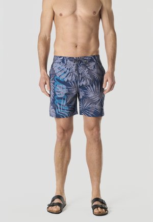 Modelo masculino vistiendo pantalones cortos de baño azules con estampado de hojas tropicales y sandalias negras de dos tiras, posando frente a un fondo claro y liso.