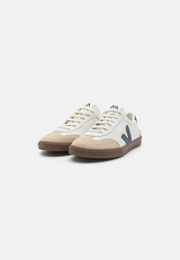 Hvite og beige sneakers med brun gummisåle. Funksjoner inkluderer en ruskinn tåhette og en blå "V"-logo på siden. Snøring.