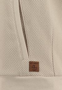 Hellbeige strukturiertes Hoodie mit einem Wabenmuster, ausgestattet mit einem Front-Reißverschluss und einem braunen Leder-Brandlabel unten links.