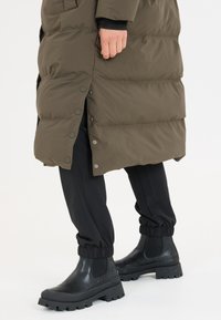 Puffiger olivgrüner Mantel mit Druckknöpfen und Seitentaschen, kombiniert mit schwarzen Hosen mit elastischen Bündchen und klobigen schwarzen Chelsea-Boots.