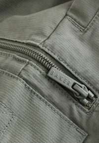 Close-up van grijze stof met een rits waarop "BAKER" is gegraveerd, benadrukt een gestructureerde afwerking en nette stiksels.