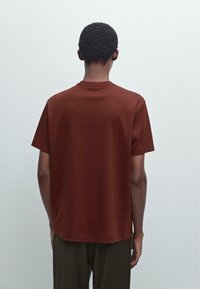 Hnědé tričko s krátkým rukávem a kulatým výstřihem, vyrobené z hladkého materiálu, se uvolněným střihem a minimalistickým designem.