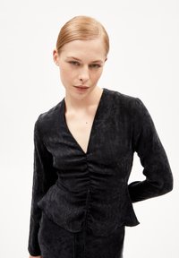 Blusa nera con maniche lunghe, scollo a V, dettagli arricciati sul davanti e orlo svasato. Tessuto morbido e motivo fantasia crea texture.