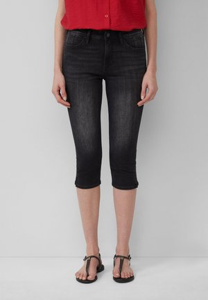 Capri en denim noir arrivant au genou avec une coupe ajustée, cinq poches et des ourlets retroussés. Associé à des sandales plates marron et une chemise rouge.