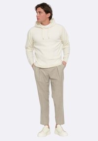 Felpa con cappuccio crema, pantaloni beige plissettati e scarpe da ginnastica bianche. Il tessuto appare morbido, con una superficie liscia e una vestibilità rilassata.