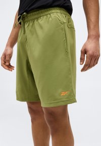 Άνδρας που φοράει σορτς Reebok σε ελιά πράσινο χρώμα με ελαστική μέση και πορτοκαλί λογότυπο κοντά στο τελείωμα, στέκεται με τα χέρια χαλαρά δίπλα στο σώμα.