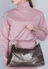 Donna in dolcevita rosa che tiene una grande borsa di pelle metallica scura con una sola tracolla davanti a uno sfondo grigio.