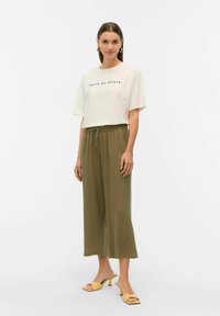 Camiseta crema con texto negro, combinada con pantalones de pierna ancha verde oliva y tacones amarillos abiertos. Conjunto simple y casual.