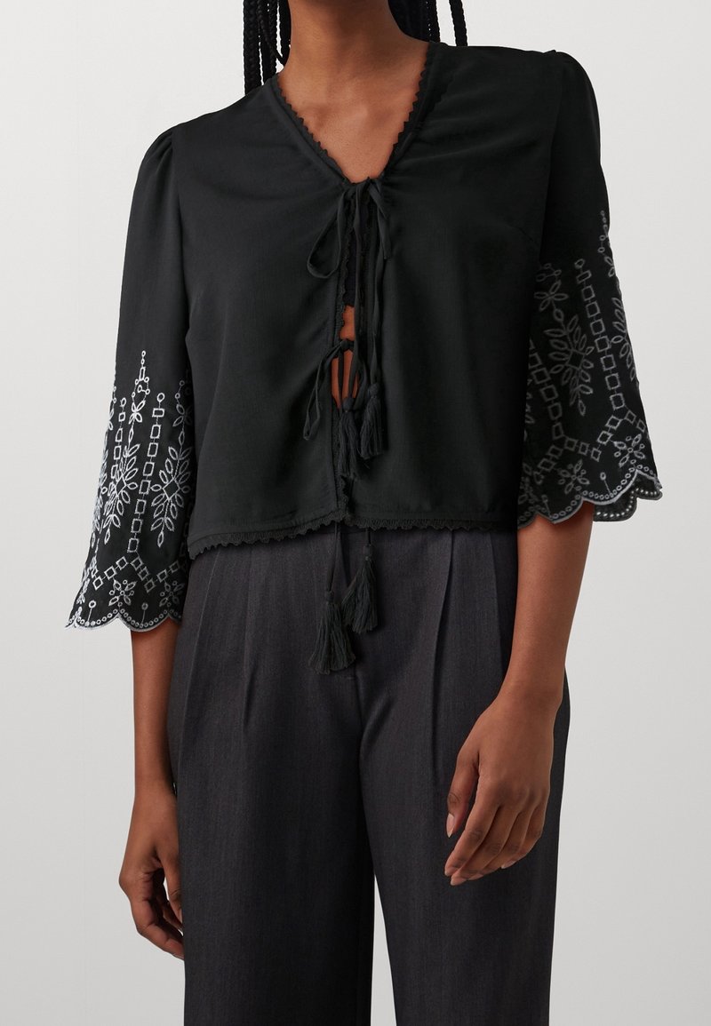 Blouse noire à col en V, nouée à l'avant, avec des manches cloche brodées présentant des motifs floraux. Accents incluant des bords festonnés et des pompons.
